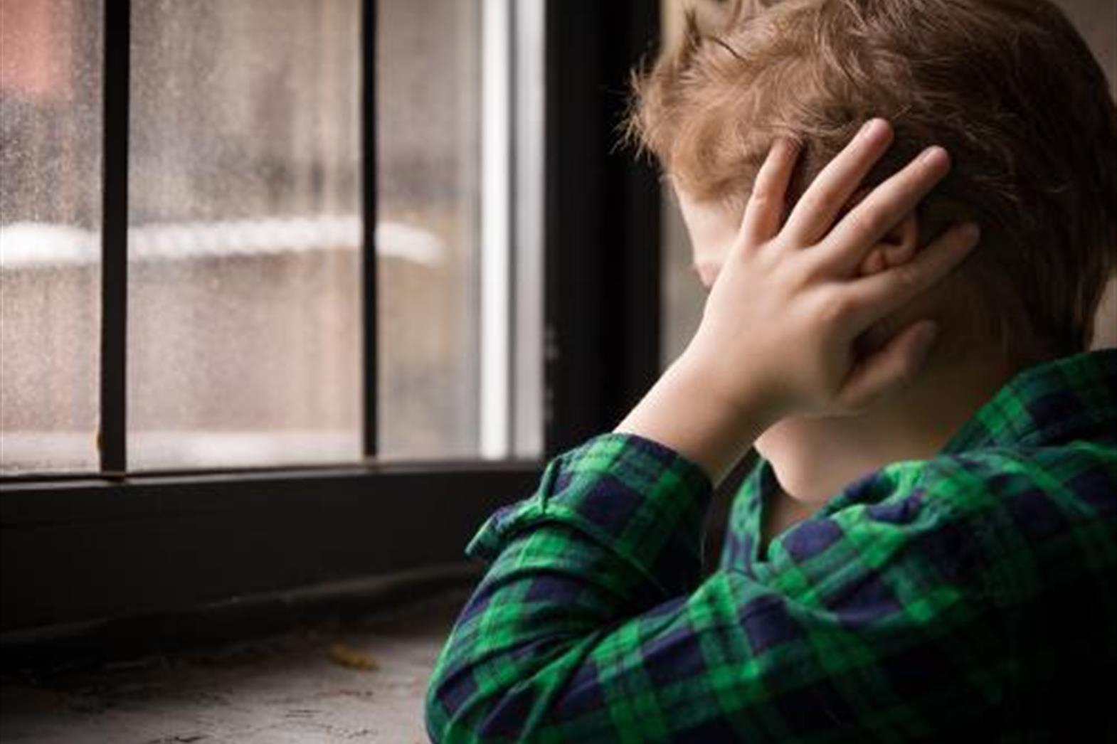A boy looking out a window and covering his ears with his hands