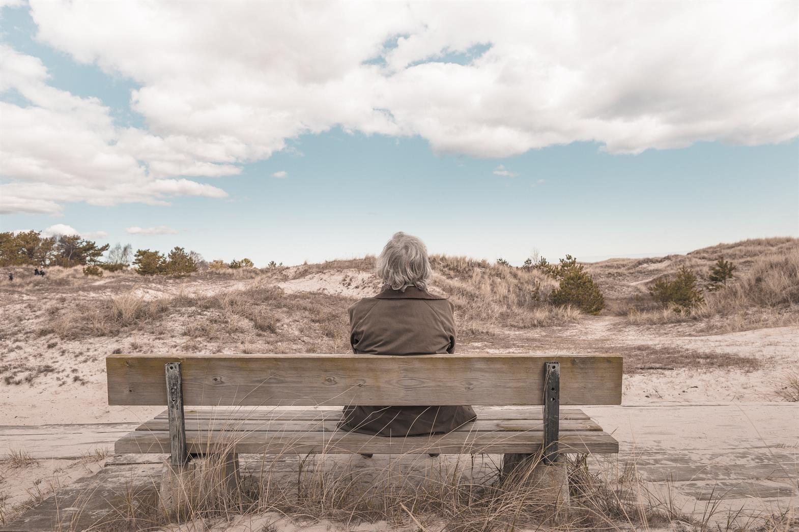 an elderly person sitting on a bench looking out over a dirt road towards the horizon. 