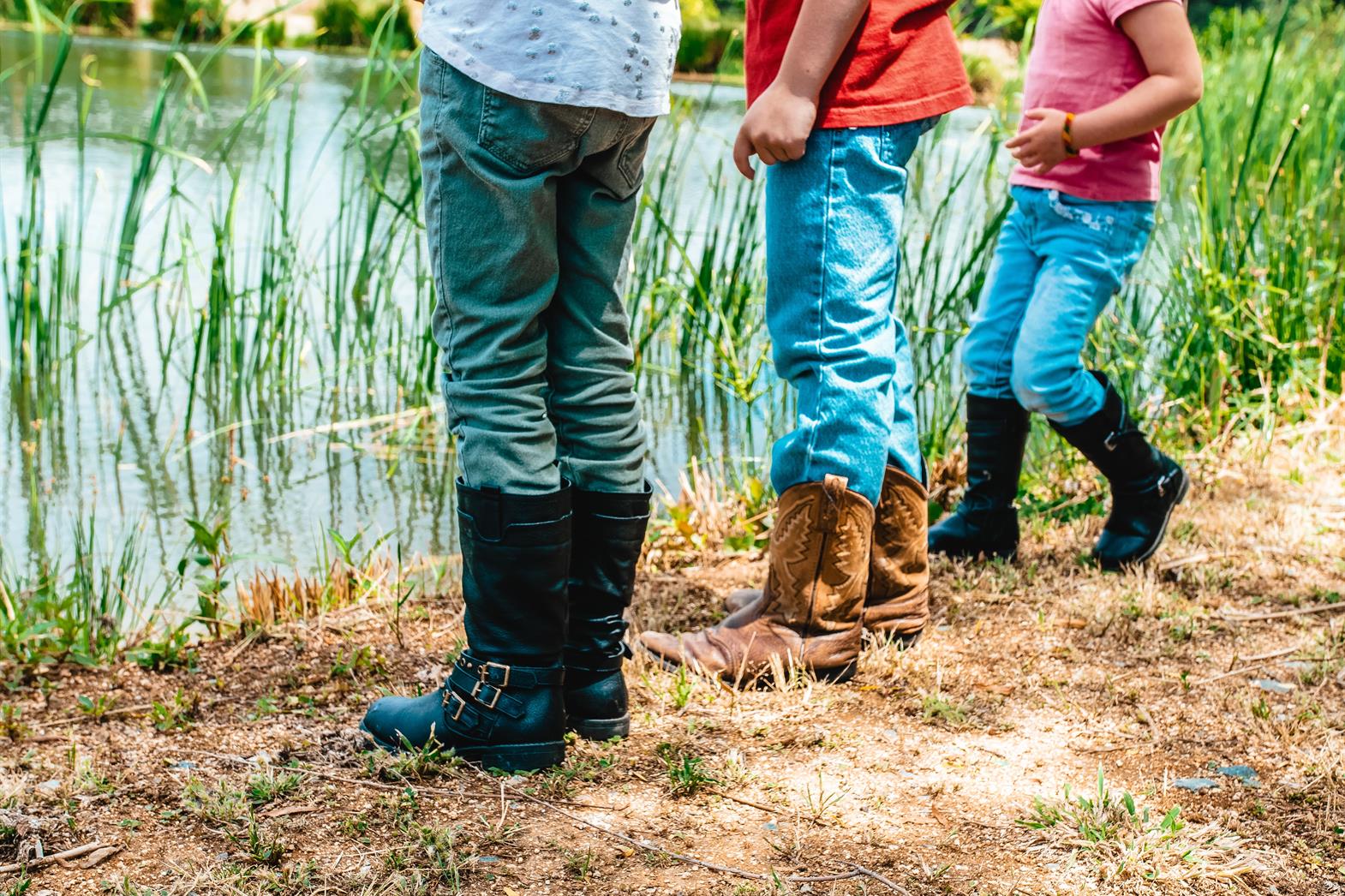 The legs of three children wearing boots and jeans and standing by a river
