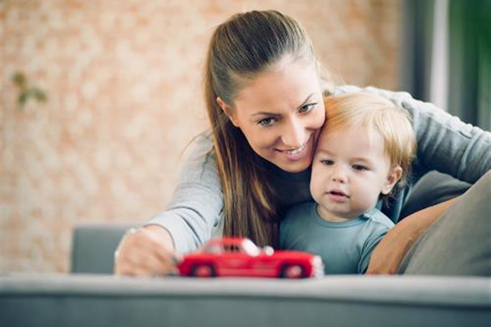 A woman in her early 20s sits with a toddler. They play with a red toy car. 